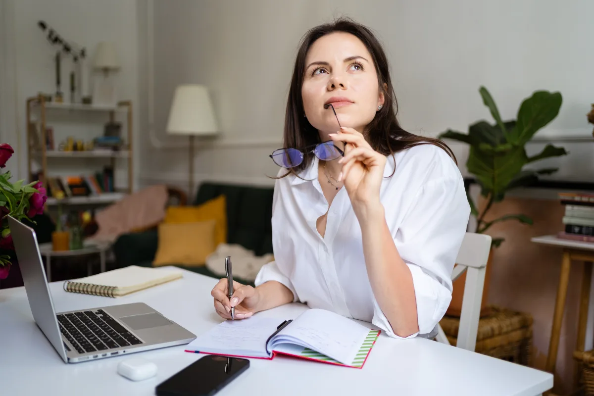 Jonge vrouw met bril in haar hand achter haar bureau kijkt nadenkend.